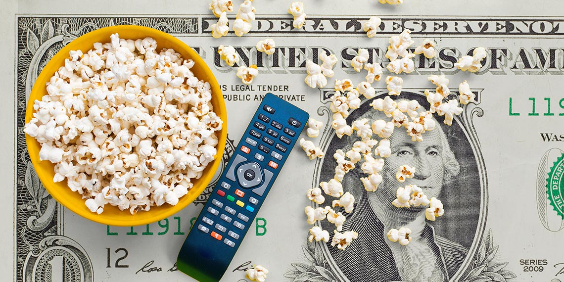 Bowl of popcorn and remote control sitting on a dollar bill table.