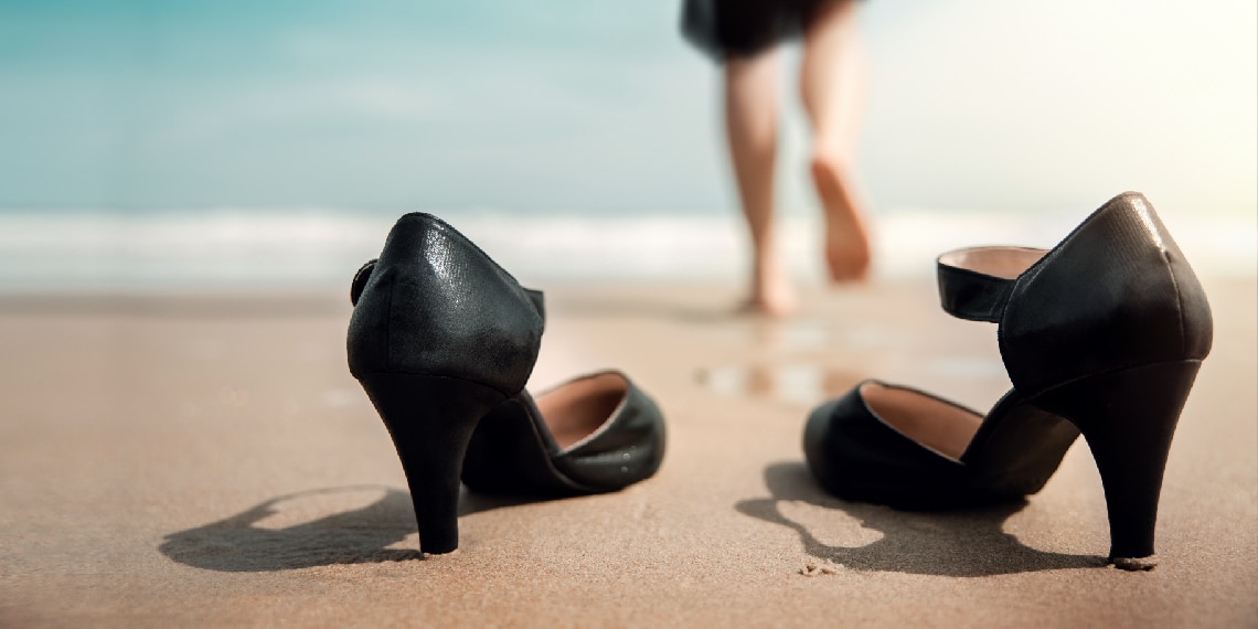 Photo of woman with business heels off in the sand as she walks towards the ocean.