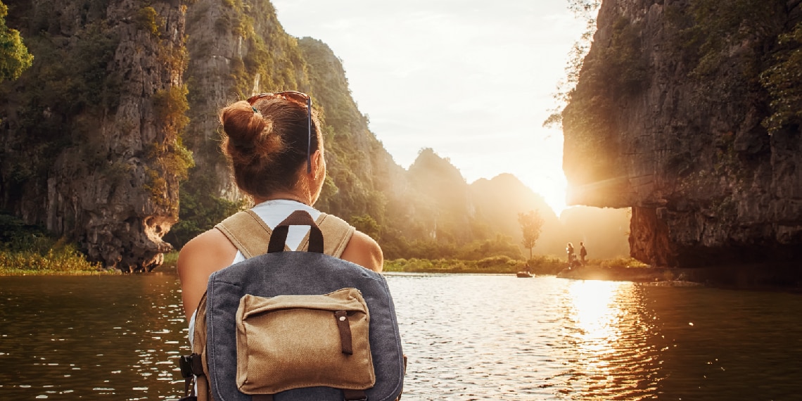 Photo of woman with backpack looking at sun hovering over water.