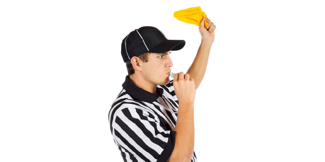 Photo of football referee blowing a whistle and holding up yellow penalty flag.